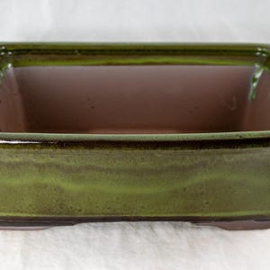 May include: A square bonsai pot with a glossy green exterior and a brown interior. The ceramic pot has a slightly flared shape and a smooth, glazed finish. The pot is designed for planting small trees.