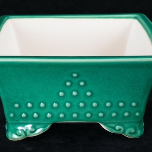May include: A square green ceramic planter with a white interior and a decorative pattern of raised dots. The planter has four feet.