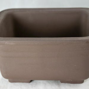 May include: A brown rectangular ceramic bonsai pot with a smooth, slightly textured surface. The pot has a wide, shallow base and four small feet.