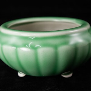 May include: A small, round, green ceramic planter with a ribbed exterior and three small white feet. The interior is a light cream color. The planter is designed for small plants or succulents and sits against a black background.