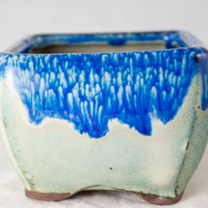 May include: Square ceramic planter with a blue and white glaze. The glaze has a mottled effect, with blue streaks and white flecks. The planter has a slightly rounded bottom and four small feet.