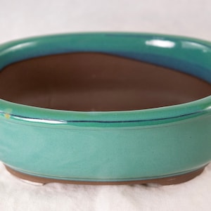 May include: A green oval ceramic bonsai pot with a brown interior. The pot has a glazed finish and a slightly raised rim.