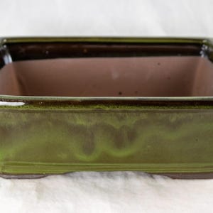 May include: A green glazed rectangular ceramic bonsai pot with a brown interior. The pot has a slightly flared rim and a rounded base.