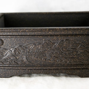 May include: A dark brown rectangular ceramic planter with a raised design of a mountain range and a wall. The planter has a textured surface and a slightly raised rim.