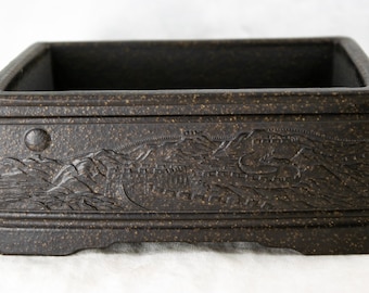 May include: A dark brown rectangular ceramic planter with a raised design of a mountain range and a wall. The planter has a textured surface and a slightly raised rim.
