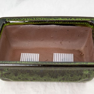 May include: A green glazed rectangular ceramic bonsai pot with two white plastic mesh squares inside.