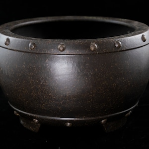 May include: A dark brown ceramic pot with a rounded shape and a wide rim. The pot has a textured surface and is decorated with small, evenly spaced studs around the rim and base. The pot has three small feet.