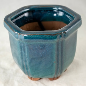 May include: A teal blue ceramic hexagonal planter with a glazed finish. The planter has a wide opening and a rounded base.