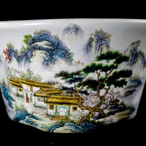 May include: White ceramic bowl with a hand-painted landscape scene. The design features mountains, trees, and traditional buildings in shades of blue, green, and brown. The bowl has a flared shape.