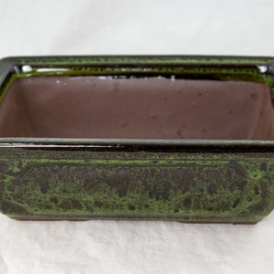 May include: A rectangular green and brown ceramic bonsai pot. The pot has a glazed finish and a slightly raised rim.