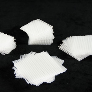 May include: Several stacks of white plastic canvas squares are arranged on a black surface. The squares have a grid-like pattern and are of varying sizes. The image is well-lit, highlighting the texture and shape of the canvas.
