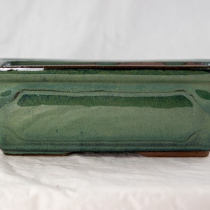 May include: A square green ceramic bonsai pot with a brown rim. The pot has a slightly textured surface and a rounded bottom.