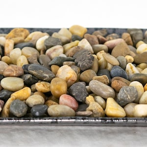 May include: A black square dish filled with a variety of smooth, rounded stones in shades of brown, gray, white, and tan.