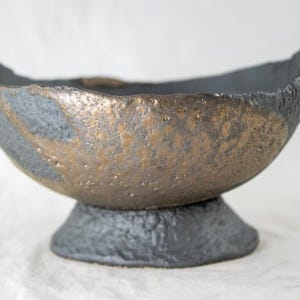 May include: A black and gold ceramic bowl with a textured surface. The bowl has a wide, rounded base and a flared lip.