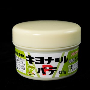 May include: A white plastic container of Kiyonaru putty, a type of modeling compound. The container is labeled in Japanese and has a red "P" on the front. The container is 135g.