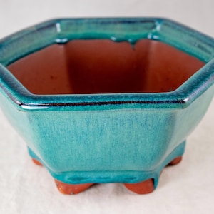 May include: A turquoise ceramic bonsai pot with a hexagonal shape. The pot has a glossy finish and a contrasting reddish-brown interior. It features three small, rounded feet and a dark blue trim around the rim.