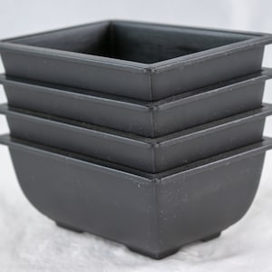 May include: Four black plastic square bonsai pots stacked on top of each other. The pots are all the same size and have a slightly rounded bottom.