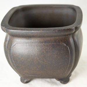 May include: A dark brown square ceramic bonsai pot with a textured surface and three small feet. The pot has a slightly rounded shape and a wide opening.