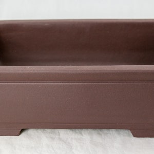 May include: A rectangular brown ceramic bonsai pot with a smooth, slightly textured surface. The pot has a wide, shallow base and a slightly flared rim.