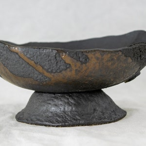 May include: A black ceramic bowl with a raised base and a brown glaze. The bowl has an irregular shape and a rough texture.