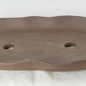 May include: A brown ceramic bonsai pot with a wavy edge and two drainage holes. The pot has a wood-grain texture.