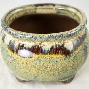 May include: A small, round ceramic planter with a speckled glaze in shades of green, brown, and cream. The planter has a wide opening, a dark brown interior, and three small feet. The rim is a darker brown.