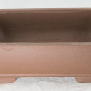 May include: A square, terracotta bonsai pot with a rounded base and four small feet. The pot is unglazed and has a rustic, earthy appearance.