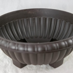 May include: A brown, round planter with a fluted interior and a decorative, ridged exterior. The planter has three short legs and a wide rim. Ideal for outdoor or indoor use.