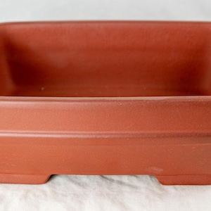 May include: A square, terracotta-colored planter with a rectangular shape. The planter has a slightly raised base with four small feet. The sides are smooth with a subtle horizontal groove near the top. Ideal for small plants or succulents.