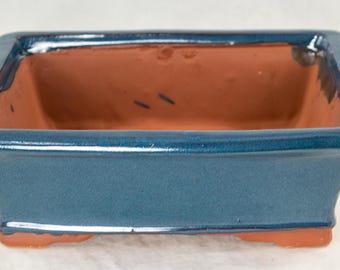 May include: A square, blue glazed bonsai pot with a terracotta interior. The pot has a slightly flared rim and a flat base.