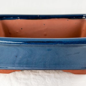 May include: A square bonsai pot with a glossy, deep blue exterior and a reddish-brown interior. The pot has a rectangular shape with slightly flared sides and small feet. The overall design is simple and elegant.