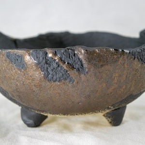May include: A small, three-legged ceramic bowl with a black and gold glaze. The bowl has an irregular, slightly wavy rim and a rough, textured surface.