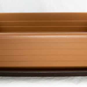 May include: A rectangular brown plastic planter with horizontal ridged detailing, sitting on a dark brown tray. The planter has a rounded rim and is designed for gardening. The overall shape is a rectangle.