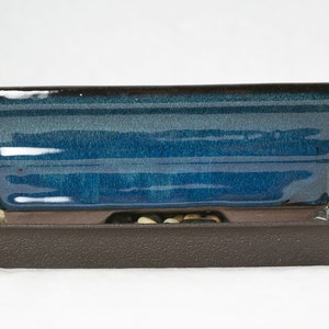 May include: A blue glazed ceramic bonsai pot with a brown plastic tray. The pot has a rectangular shape with rounded edges and a slightly flared lip. The tray has a textured surface and is designed to catch excess water.