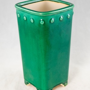 May include: A green ceramic planter with a square shape and a slightly flared top. The planter has a decorative rim with small raised dots and a geometric pattern around the base.