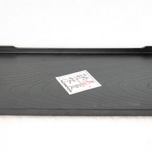May include: A black rectangular serving tray with a wood grain pattern. The tray has slightly raised edges and a small white label with Japanese characters on it.