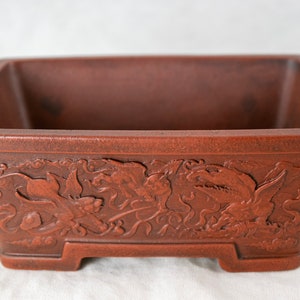 May include: A rectangular terracotta bonsai pot with a decorative design of stylized flowers and leaves. The pot has a brown glaze and a slightly textured surface.