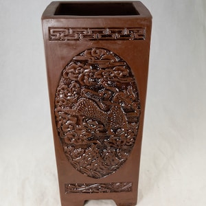 May include: A brown ceramic planter with a raised design of a dragon and clouds. The planter has a square shape with a rectangular opening at the top.