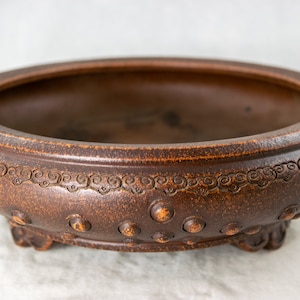 May include: A brown ceramic bonsai pot with a rounded shape and a decorative design of swirling lines around the rim. The pot has a series of small, round bumps around the base and three small feet.