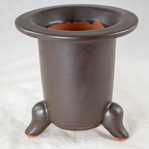 May include: A brown ceramic planter with a wide brim and three curved feet. The interior is a contrasting terracotta color. The planter has a cylindrical body and a smooth, matte finish. The feet have orange tips.