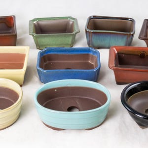 May include: Assortment of ten ceramic bonsai pots in various colors and shapes, including square, rectangular, and oval. The pots are glazed and have a variety of colors, including brown, green, blue, and yellow.