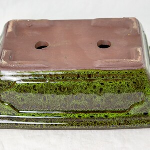 May include: A square ceramic bonsai pot with a green glaze and brown accents. The pot has two drainage holes on the bottom.