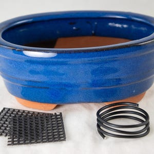 May include: A blue oval ceramic bonsai pot with a drainage hole and a wire grid for drainage. The pot is sitting on a white surface with a black wire coil next to it.