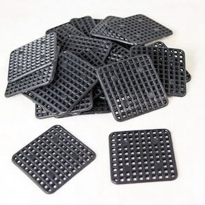 May include: A pile of black, square plastic mesh sheets with a grid pattern. Several sheets are stacked together, with a few lying separately. The sheets have rounded corners and small raised dots on the back.
