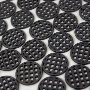 May include: A close-up of a large quantity of black plastic circular mats with a grid pattern. The mats are arranged in a random pattern on a white surface.