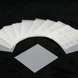 May include: A collection of white plastic canvas sheets arranged on a black surface. The sheets are square with a grid pattern of evenly spaced holes, ideal for needlepoint and cross-stitch projects. The canvas is a bright white color.
