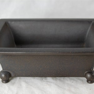 May include: A dark brown rectangular ceramic bonsai pot with four rounded feet.