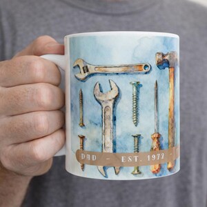 May include: White ceramic mug featuring a watercolor design of tools including a wrench, hammer, and screwdriver against a light blue background. The mug has the text "DAD - EST. 1972" on a brown banner.