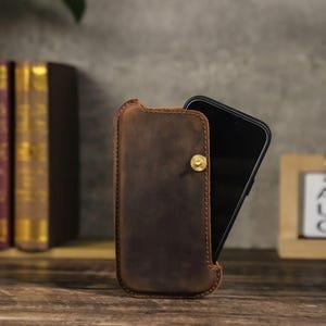 May include: A brown leather phone case with a gold-colored button closure. The case is open, revealing a black smartphone. The case is made of textured leather and has stitched edges. The background includes books and a wooden shelf.