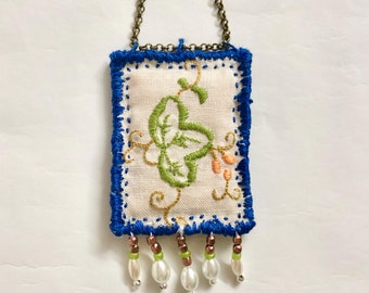 May include: A white fabric pendant with blue trim and a green embroidered floral design. The pendant is attached to a gold chain and has a beaded fringe with white and green beads.